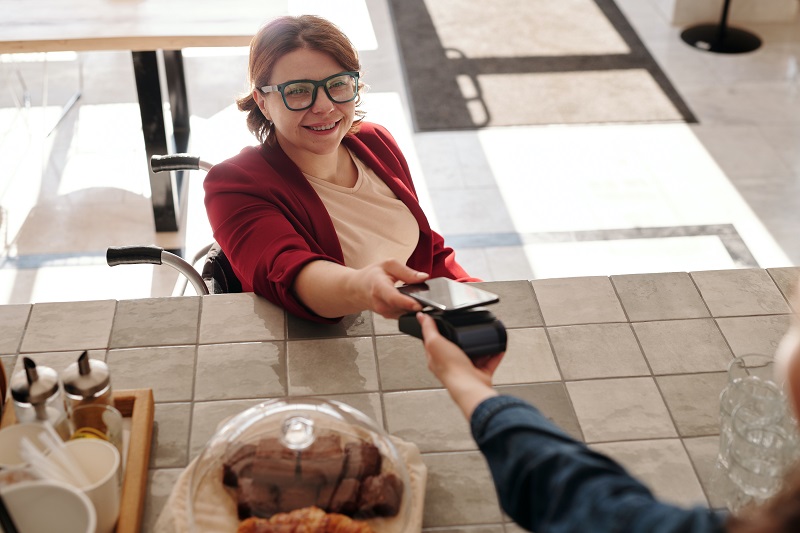 A woman in a wheelchair at a counter, smiling while making a mobile payment with her smartphone.