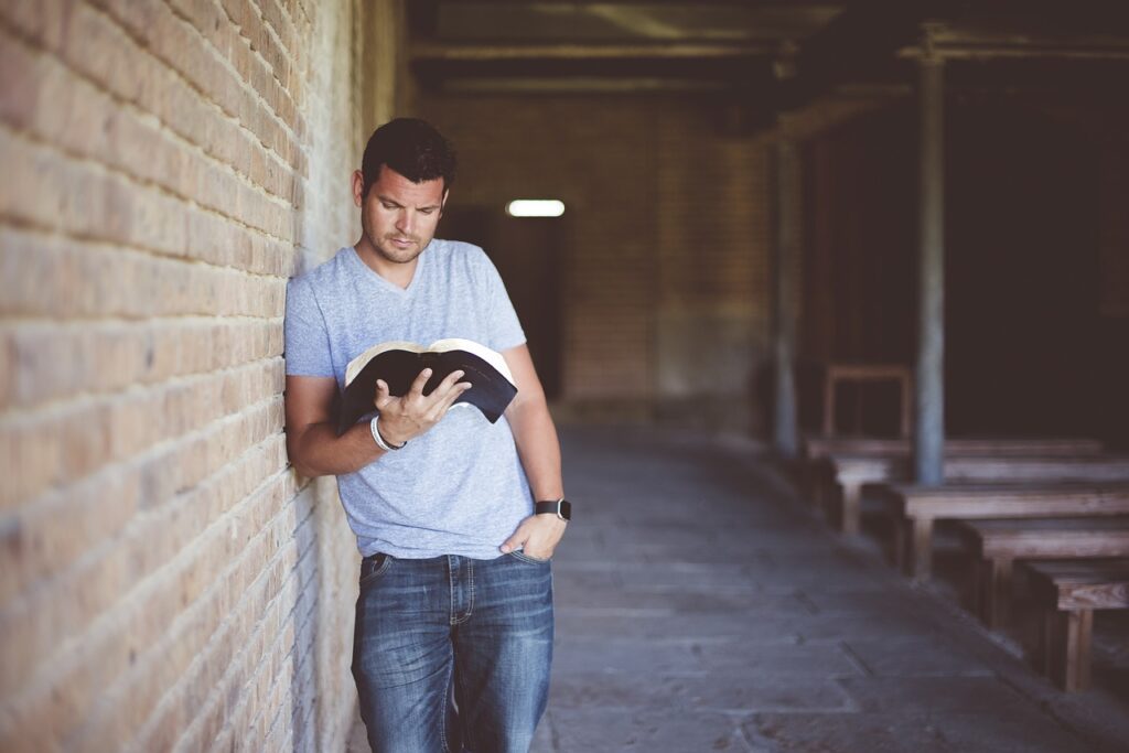 A man in a light blue t-shirt and jeans stands leaning against a brick wall while studying a book on Japanese hiragana and katakana in a quiet, rustic setting.