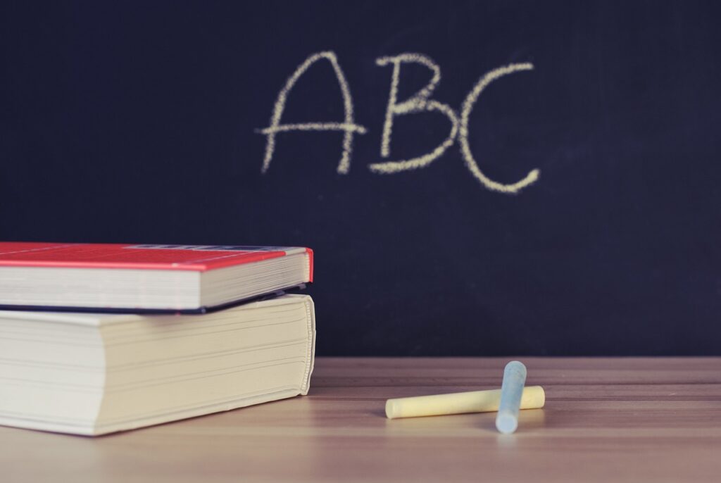 Two books stacked on a table with chalk sticks, with "ABC" written on a chalkboard in the background.