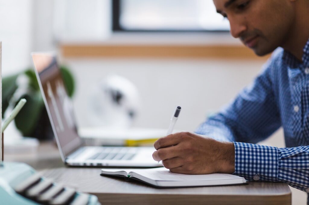 A person writing notes at a desk with a pen and notebook, while a laptop is open, representing focused study habits for learning Japanese.