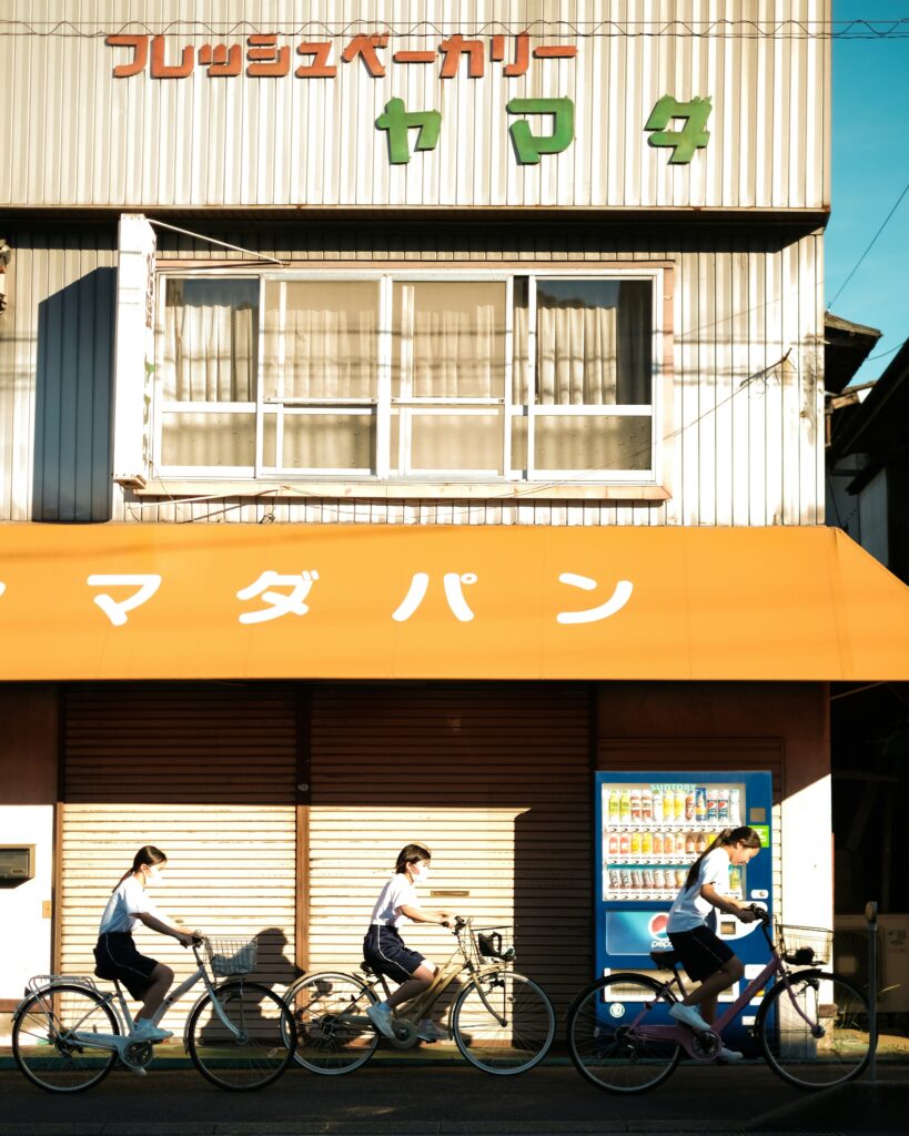 Three students riding bicycles past a bakery with a sign in Japanese characters.