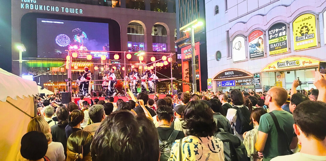 A lively crowd gathered in front of the Tokyu Kabukicho Tower during a bon odori festival, with performers on stage and a large screen displaying fireworks in the background.