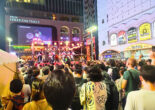 A lively crowd gathered in front of the Tokyu Kabukicho Tower during a bon odori festival, with performers on stage and a large screen displaying fireworks in the background.