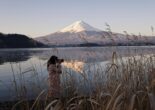 A person taking a photo of Mount Fuji by the lake with reeds in the foreground.