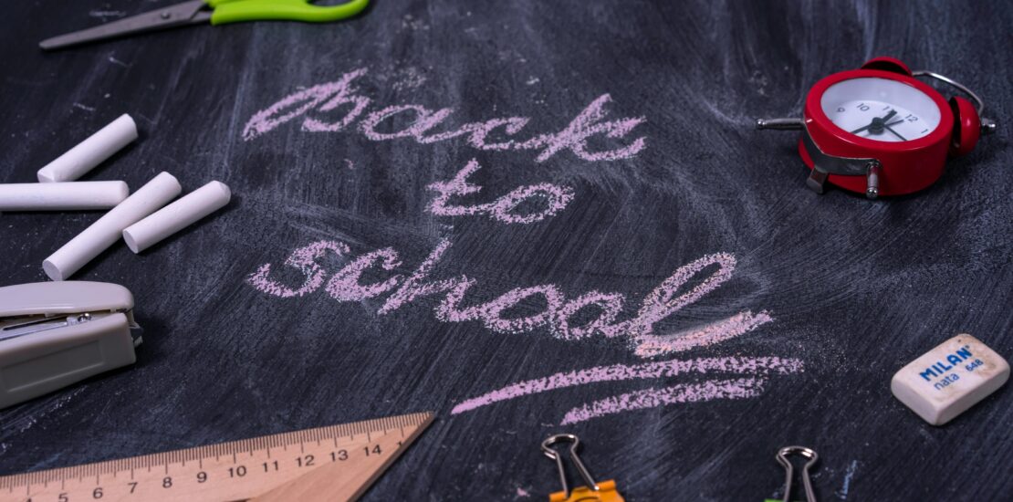 The words "Back to School" written in pink chalk on a black chalkboard, surrounded by school supplies such as chalk, scissors, and a ruler.