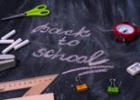 The words "Back to School" written in pink chalk on a black chalkboard, surrounded by school supplies such as chalk, scissors, and a ruler.