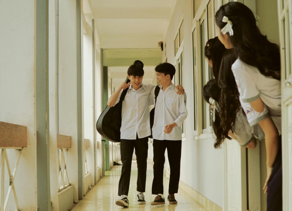 Two Japanese male students walking down a school hallway, smiling and talking, with one carrying a guitar case on his back. Female students peek around the corner, watching them.