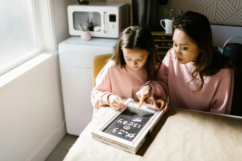 A Japanese mother and her daughter sitting at a table, working on homework together with a small chalkboard in front of them.