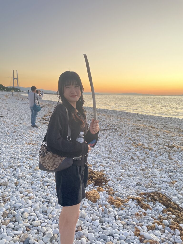 A young woman holding a stick on a pebble beach during sunset near Rinku Town. 