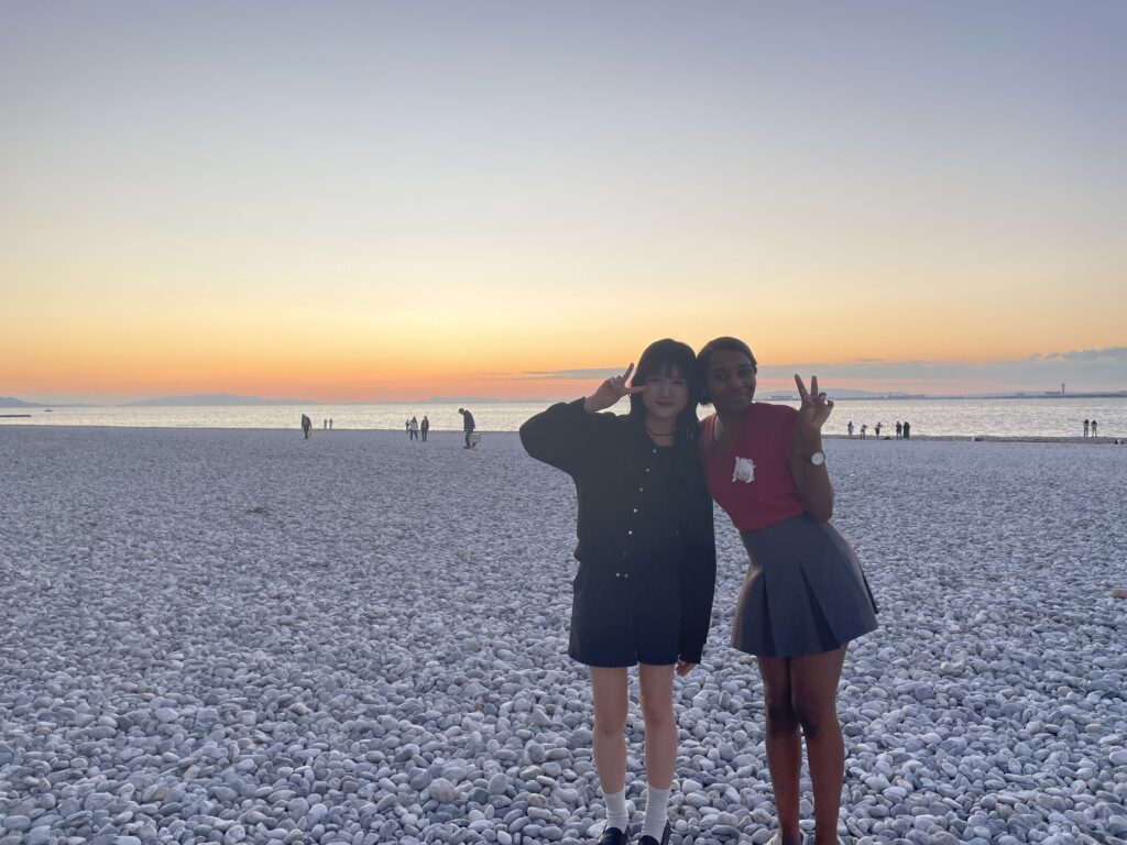 Two women posing with peace signs on a pebble beach during sunset at Rinku Town. 