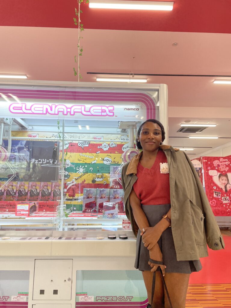 A woman posing in front of a UFO catcher at a game center in Rinku Town. Title: Fun at Rinku Town Game Center