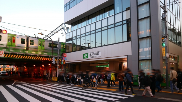 Shin-Okubo Station entrance with people walking by and a Yamanote Line train passing overhead. Shin-Okubo