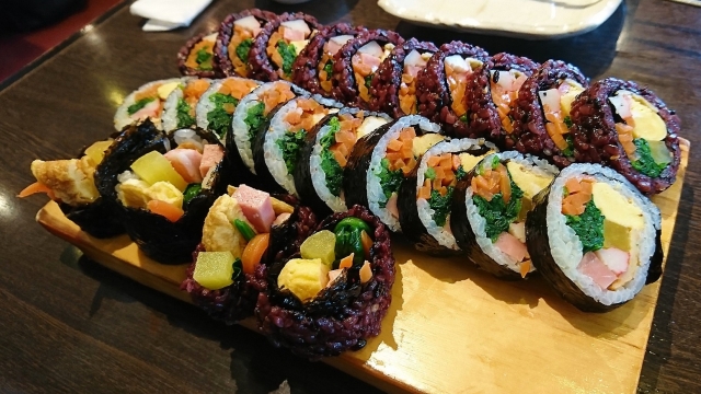 Colorful rolls of kimbap arranged on a wooden platter, featuring vegetables, pickles, and egg in Shin-Okubo.