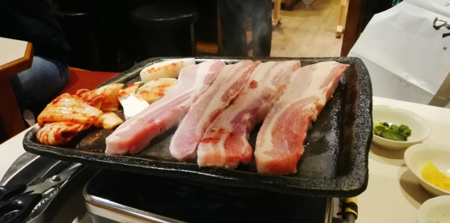 Sizzling strips of pork belly cooking on a grill, with kimchi and vegetables on the side in Shin-Okubo.