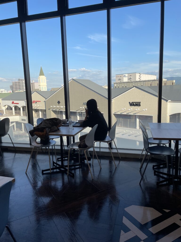 View from inside a cafe with a person looking out at the outlet stores outside. Title: Scenic Cafe View at Rinku Town