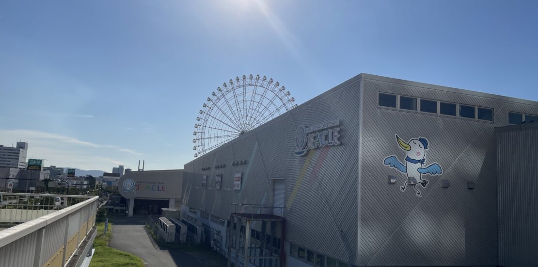 Exterior of Rinku Pleasure Town Seacle with a Ferris wheel in the background under a bright sun. Title: Seacle at Rinku Pleasure Town on a Sunny Day