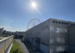 Exterior of Rinku Pleasure Town Seacle with a Ferris wheel in the background under a bright sun. Title: Seacle at Rinku Pleasure Town on a Sunny Day