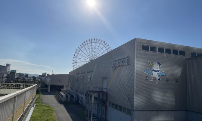 Exterior of Rinku Pleasure Town Seacle with a Ferris wheel in the background under a bright sun. Title: Seacle at Rinku Pleasure Town on a Sunny Day