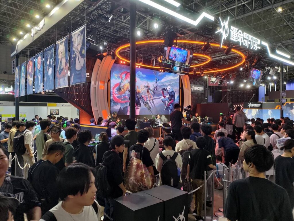 Large crowd gathered at a vibrant gaming booth showcasing banners and a dynamic game demo at TGS 2024.