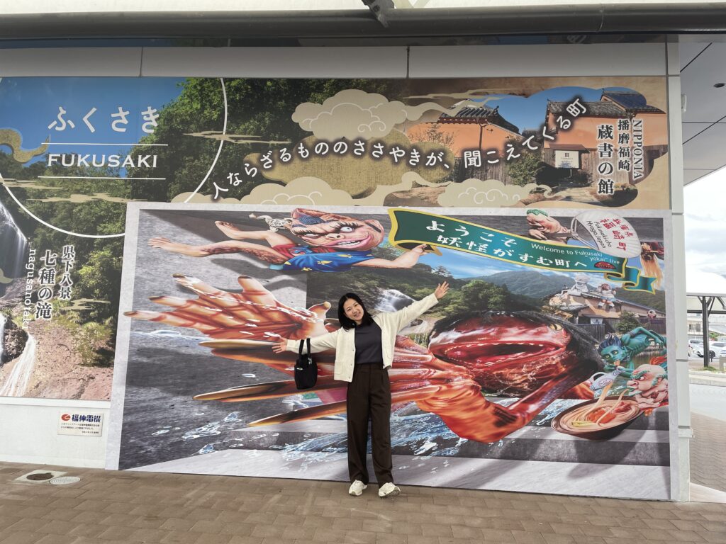 A visitor standing in front of a colorful “Welcome to Fukusaki” sign featuring images of Japanese folklore creatures.