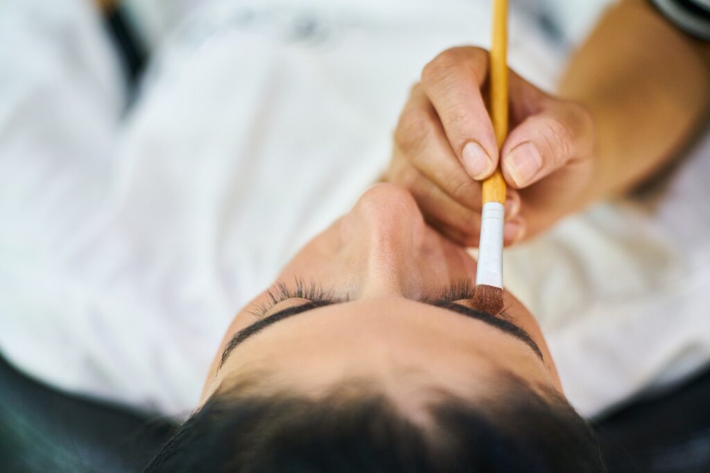 A close-up of a beauty technician applying makeup or preparing for an eyebrow or eyelash procedure on a client lying down.