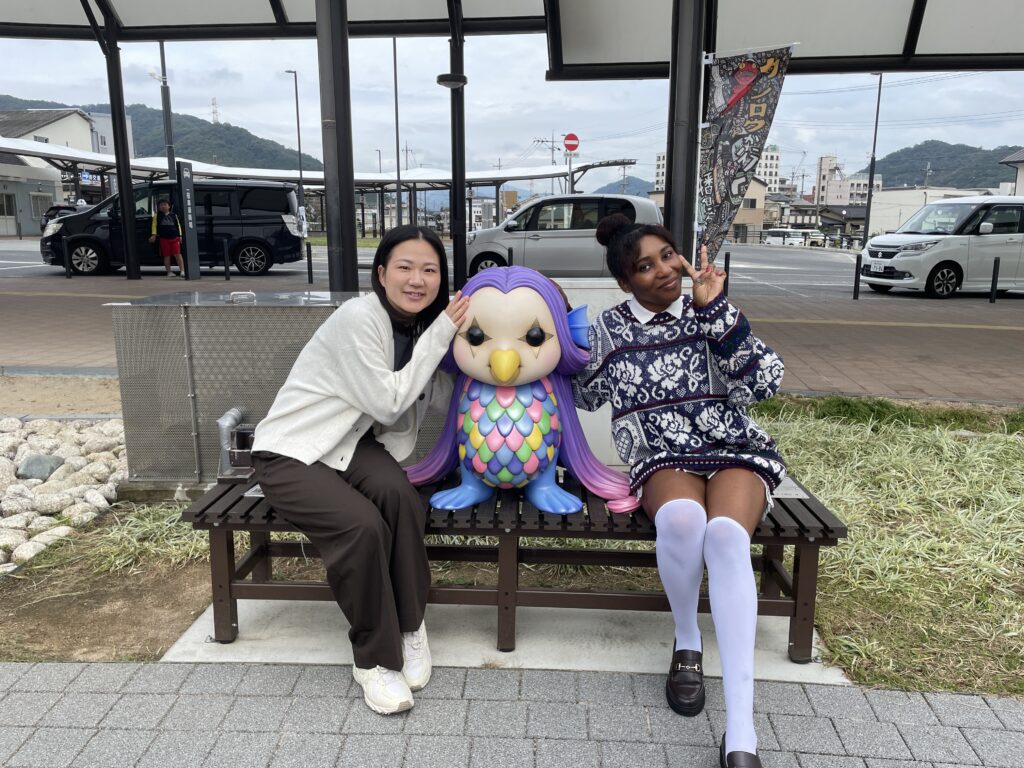 Two women sitting on a bench, posing with a colorful owl-like demon statue in Fukusaki City.