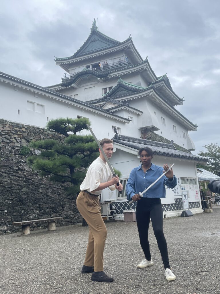 Two tourists hold practice swords while posing playfully in front of Wakayama Castle, enjoying the castle’s ninja experience.