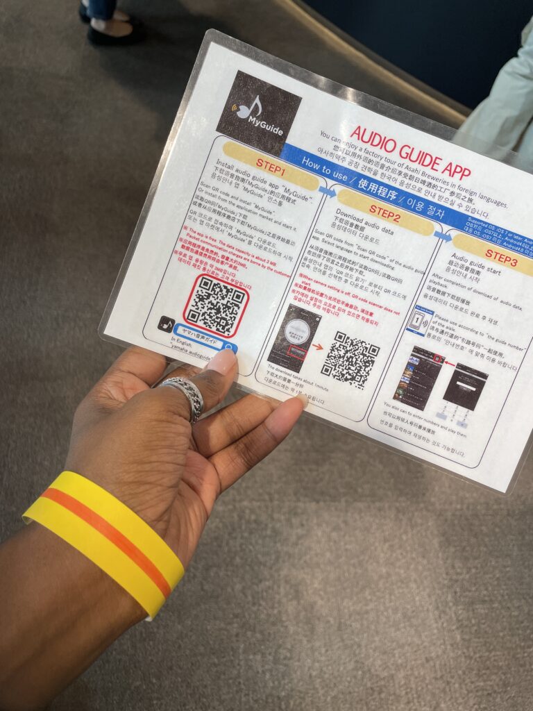 A hand holding an Asahi Beer Museum bracelet and an audio guide instruction sheet.