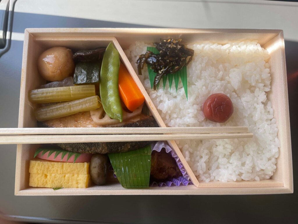 A traditional Japanese bento box featuring rice, vegetables, tamagoyaki, and a plum, with a pair of wooden chopsticks on top, enjoyed during the journey to the festival.