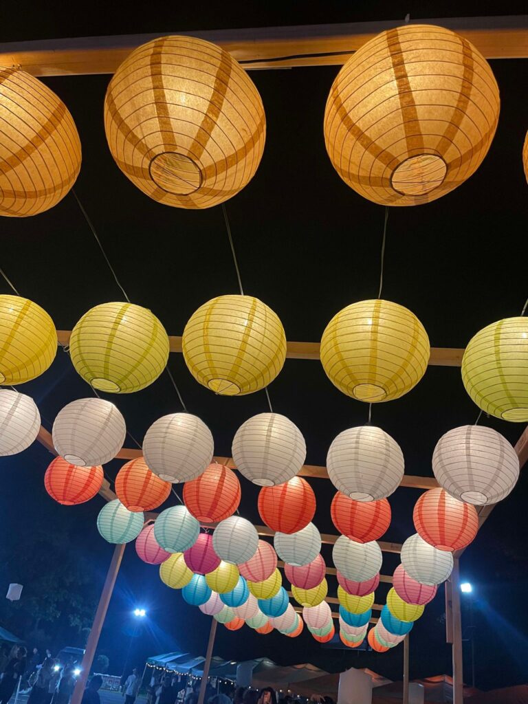 Rows of hanging paper lanterns in pastel colors, including yellow, pink, and blue, are illuminated at night, casting a festive glow over the festival venue.