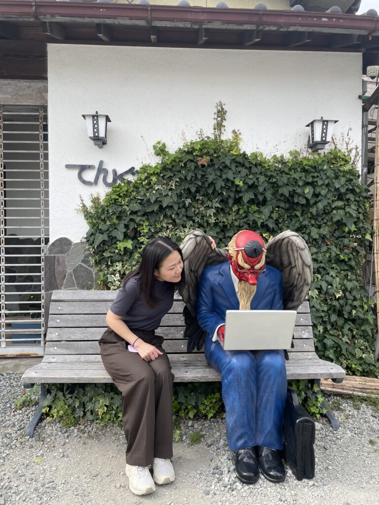 A woman sits on a bench next to a demon statue with angel wings, engrossed in a laptop.