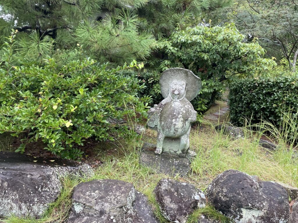 A tanuki (Japanese raccoon dog) statue with a large hat stands in a garden area, surrounded by greenery.