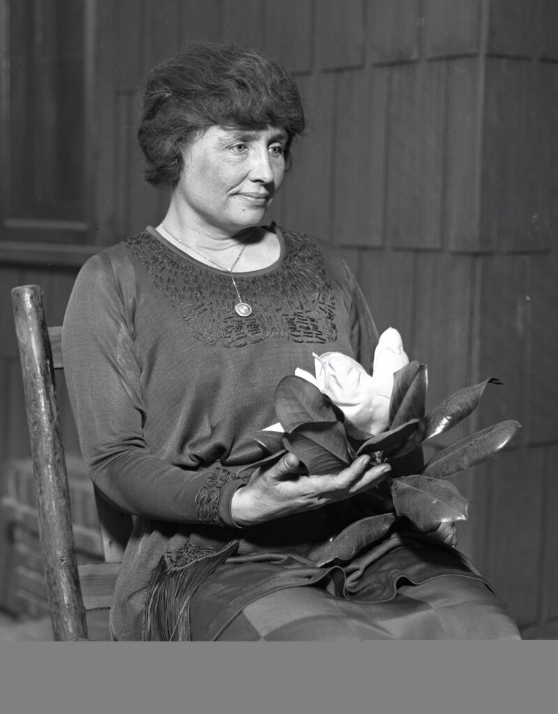 A historical photograph of Helen Keller holding a flower, dressed in a formal outfit.