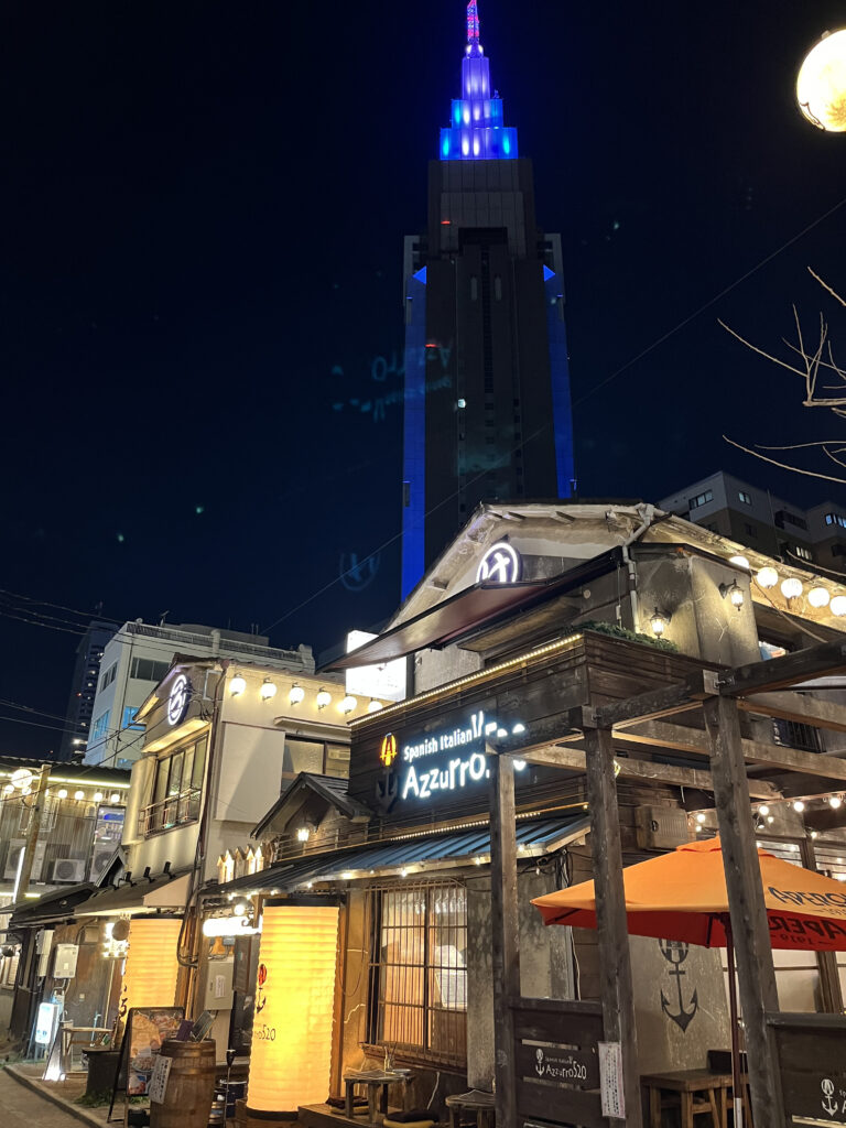 Night view of Yoyogi restaurant district with a modern skyscraper illuminated in blue in the background.