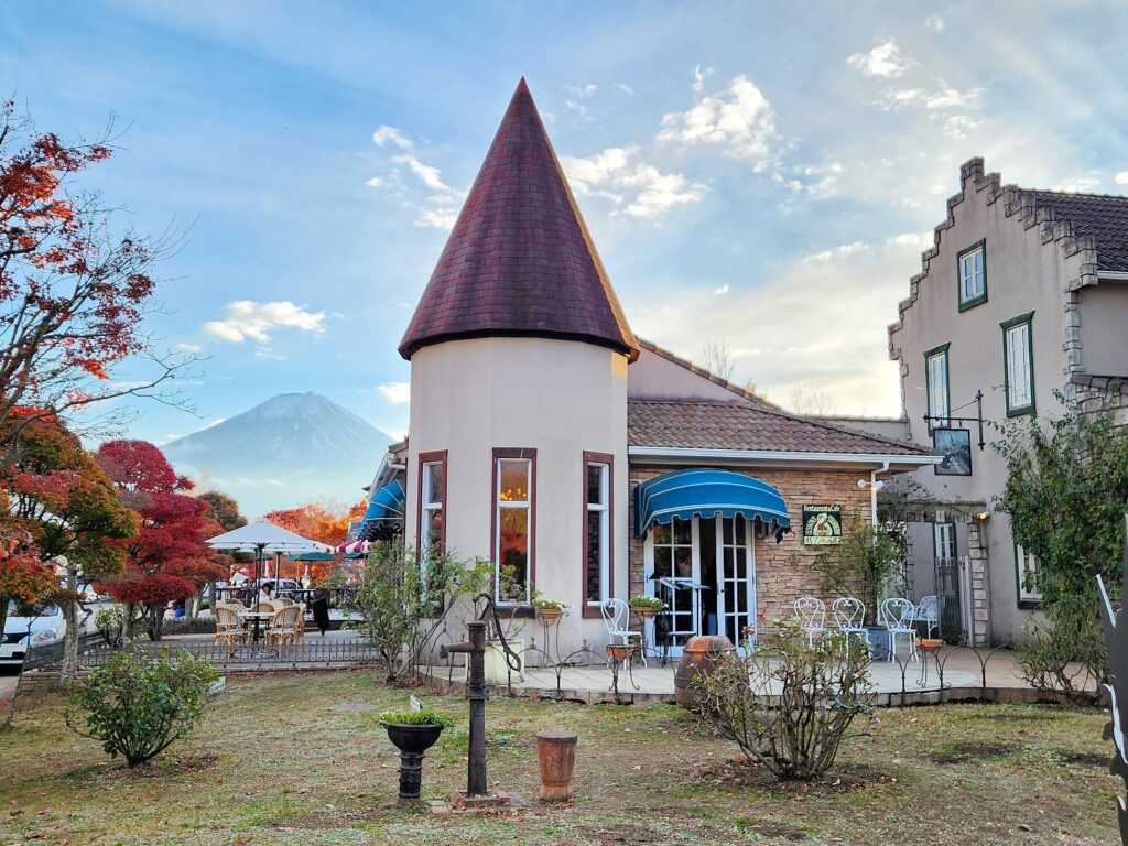 A picturesque European-style building at Kawaguchiko Konohana Museum, with Mt. Fuji visible in the background surrounded by autumn trees.