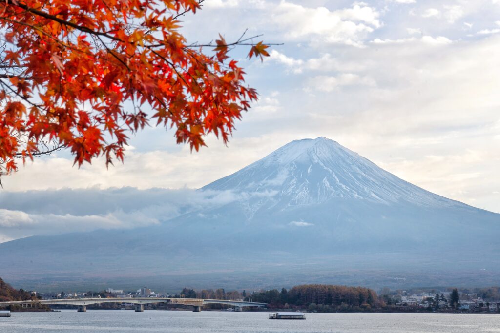 Mt. Fuji framed by vibrant red maple leaves, with Lake Kawaguchi and a bridge in the foreground under a partly cloudy sky.