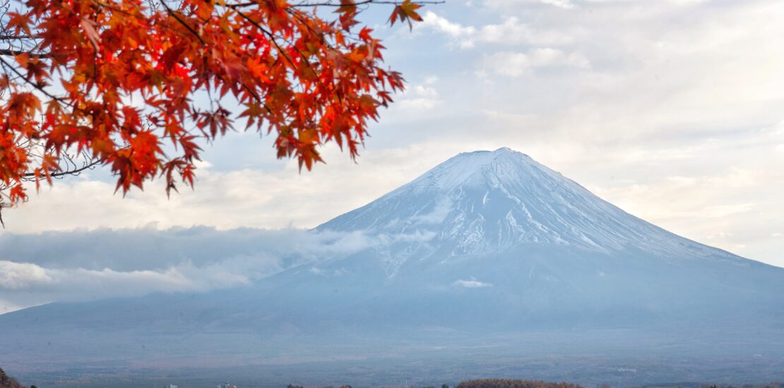 Mt. Fuji framed by vibrant red maple leaves, with Lake Kawaguchi and a bridge in the foreground under a partly cloudy sky.