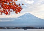 Mt. Fuji framed by vibrant red maple leaves, with Lake Kawaguchi and a bridge in the foreground under a partly cloudy sky.