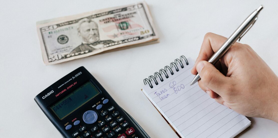 A person writing in a notepad with a calculator and cash on the table, budgeting finances and tracking savings in Japan.