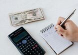 A person writing in a notepad with a calculator and cash on the table, budgeting finances and tracking savings in Japan.