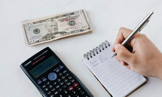 A person writing in a notepad with a calculator and cash on the table, budgeting finances and tracking savings in Japan.