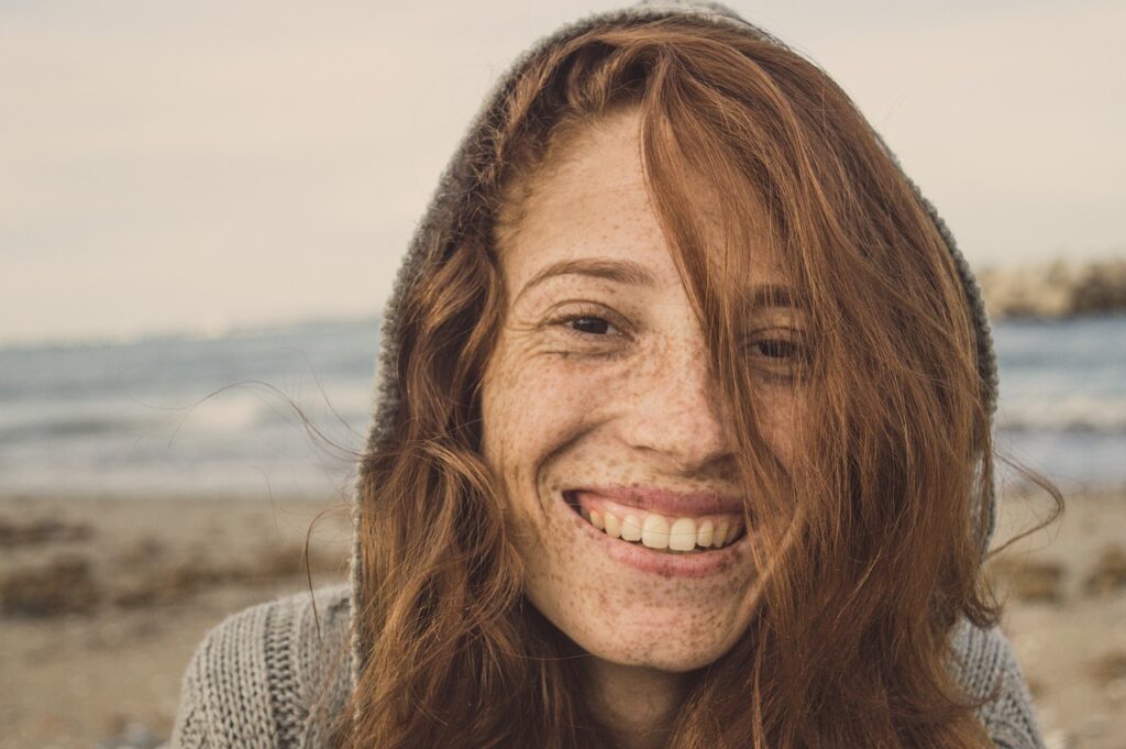 A cheerful woman smiling brightly, showcasing her healthy, white teeth against a serene beach backdrop.