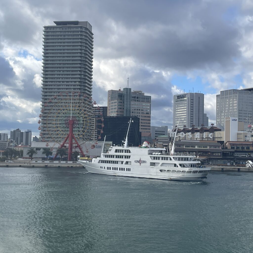 A white cruise ship in a harbor with a Ferris wheel and high-rise buildings in the background under a cloudy sky.