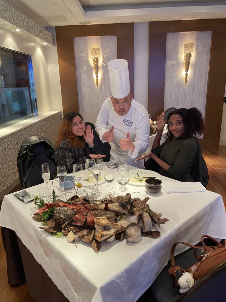 A chef wearing a tall white hat posing with two guests at a table filled with decorative elements and wine glasses.