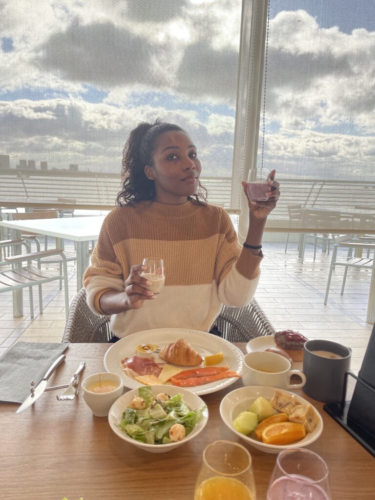 A woman enjoying a breakfast spread featuring croissants, fresh fruits, and various international dishes, with a cloudy sky visible through the large windows.