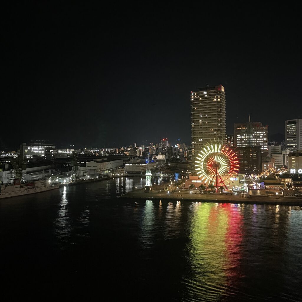 A Ferris wheel and high-rise buildings illuminated at night, reflecting on the calm waters of the harbor.