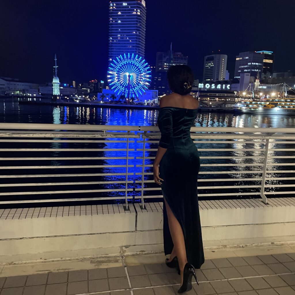 A woman in a green gown gazing at the illuminated Ferris wheel and cityscape from a waterfront balcony.