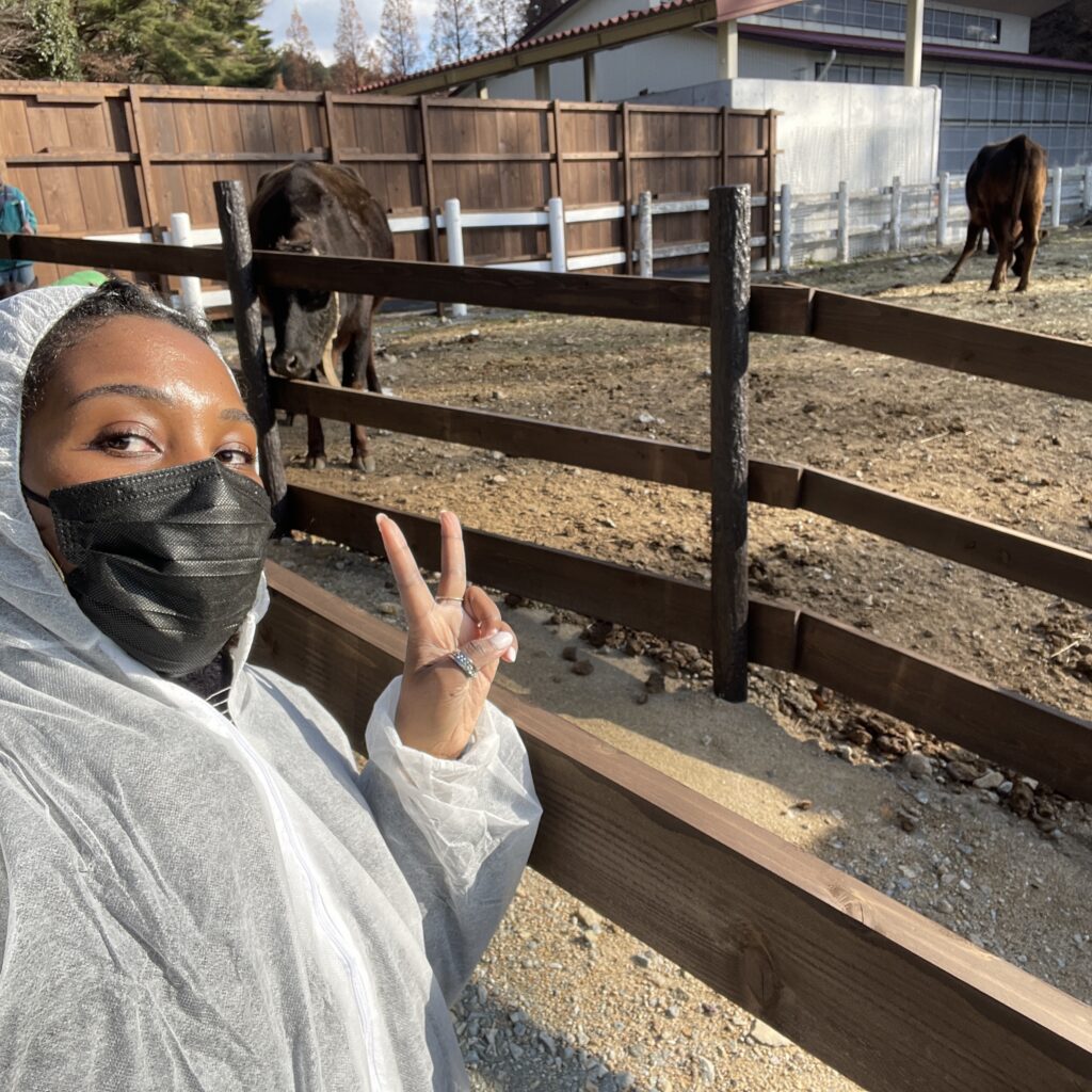 A woman in a white hazmat suit posing near a fence with cattle grazing in the background.