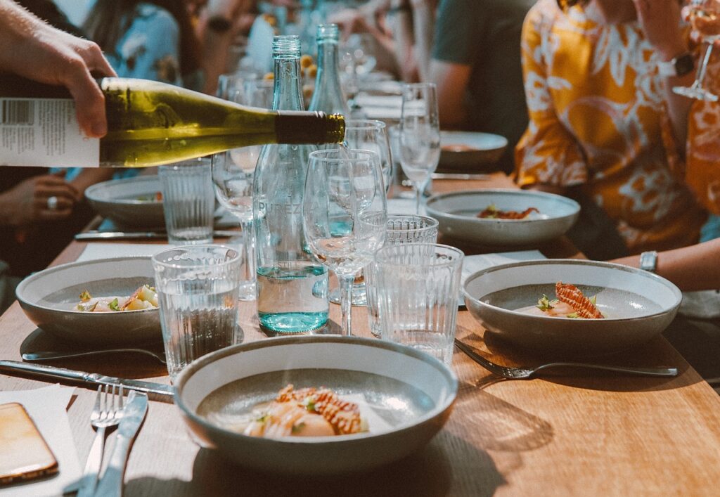 A rustic dining table set with wine, glasses, and plates, with diners visible in the background.