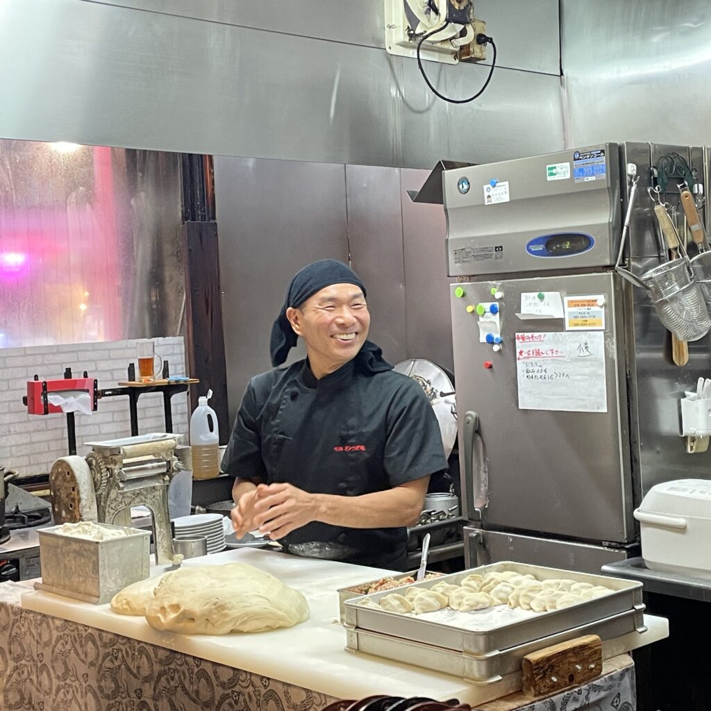 A smiling chef wearing a black bandana and apron, preparing gyoza dumplings in a kitchen.